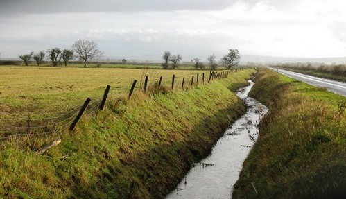 drainage ditch along side a road and farm field with a wooden and wire fence